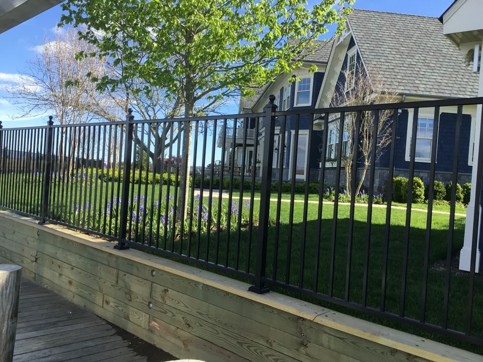 Black aluminum fence mounted along a raised timber edge with a blue custom home and lawn beyond in East Islip, New York