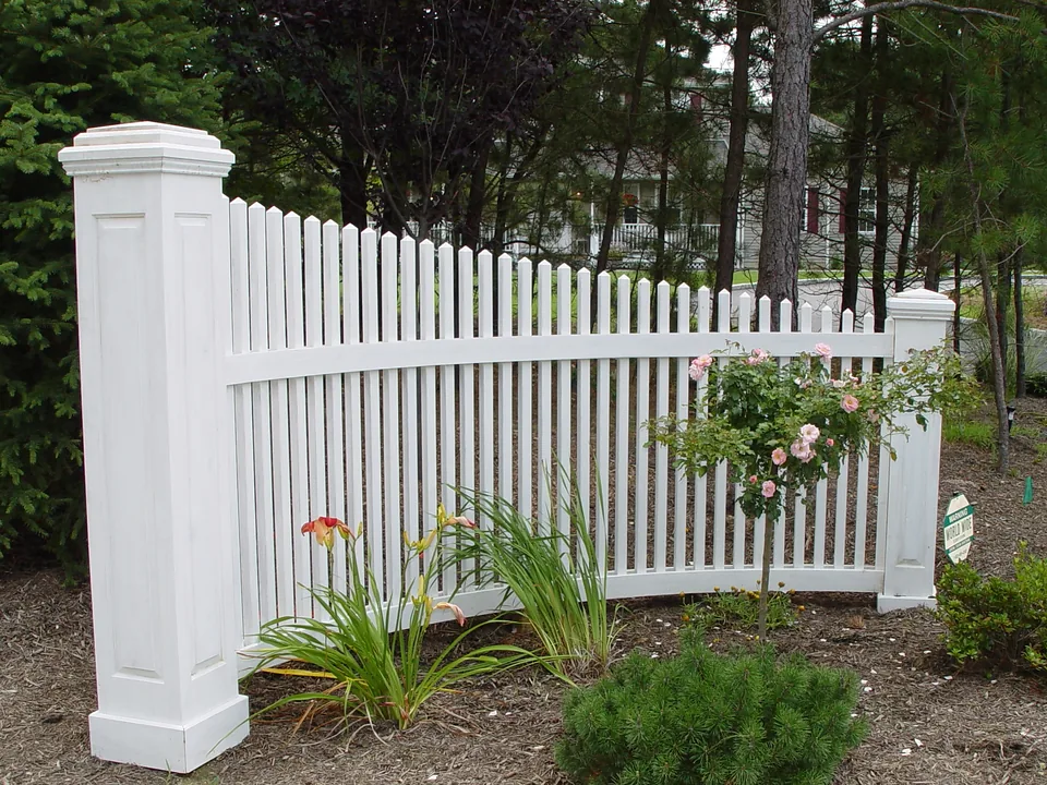 Curved cedar picket driveway entrance accent stained white, with heavy raised-panel posts, set in a mulched planting bed in Manorville, New York