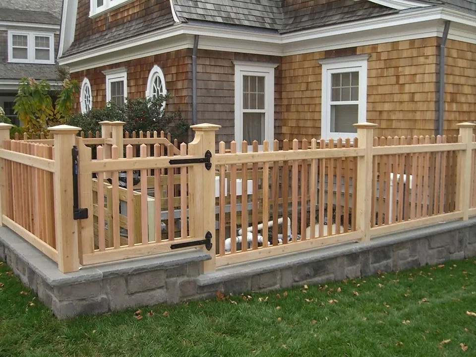 Custom-built cedar pool filter enclosure about three feet tall on a bluestone-capped stone wall, with a matching gate and black strap hinges, beside a shingled house in Oyster Bay, New York