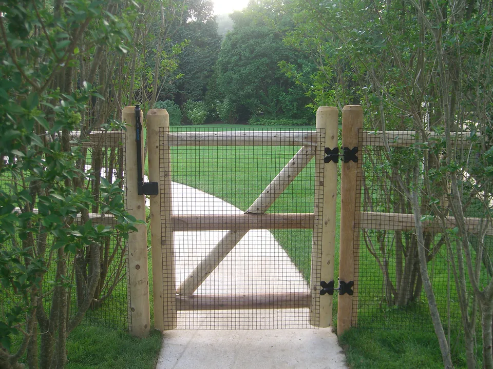 Cedar post and rail gate with an X brace and black welded wire backing, hung on peeled cedar posts with self-closing hardware, in Southampton, New York