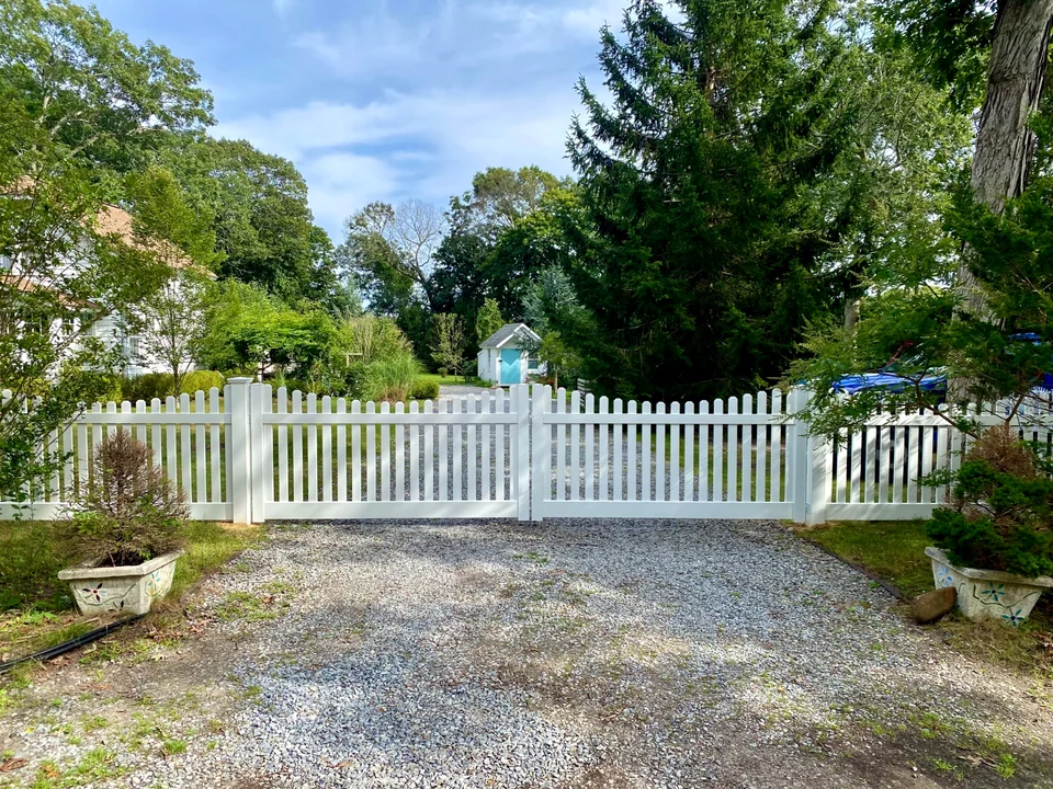 Wide driveway view of a white PVC picket double gate and fence in Speonk, New York.
